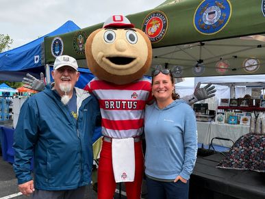 Two people posing with Ohio State's Brutus Buckeye mascot at an outdoor event.