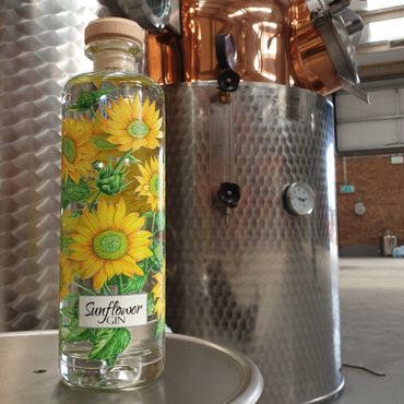 A bottle of sunflower gin with bright sunflower illustrations in front of distillery equipment.