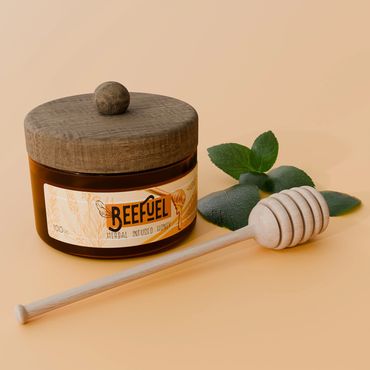 A jar of BeeFuel herbal infused honey with a wooden honey dipper and green leaves.