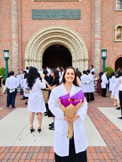 Smiling woman in white coat holding flowers outside university building.