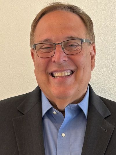 Smiling man in glasses wearing a blue shirt and dark blazer.