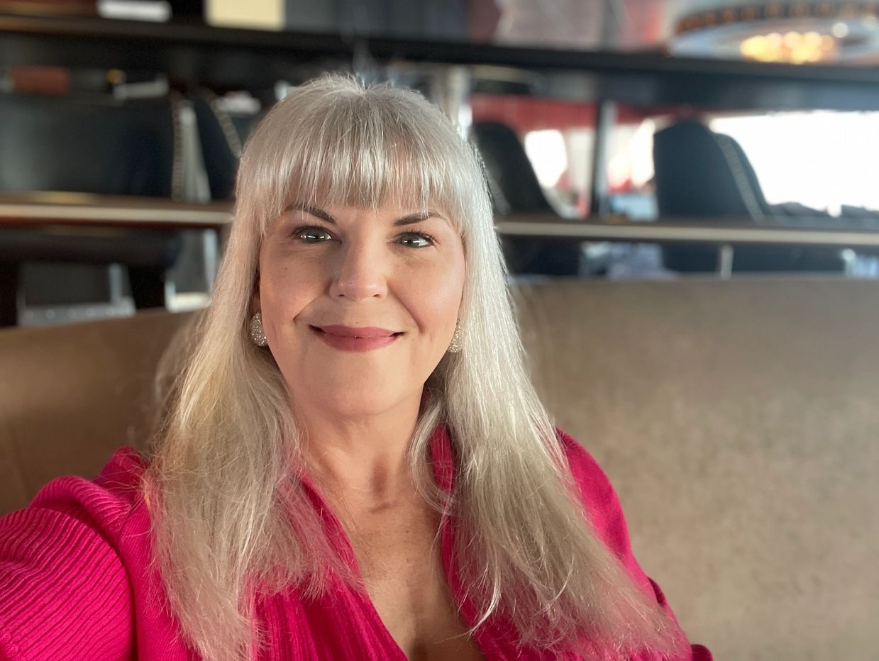 Smiling woman with long silver hair wearing a bright pink top indoors.