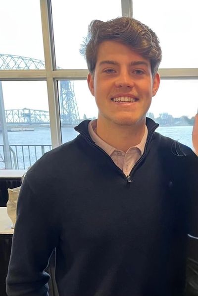 A young man smiling indoors with a waterfront and bridge visible through the window.