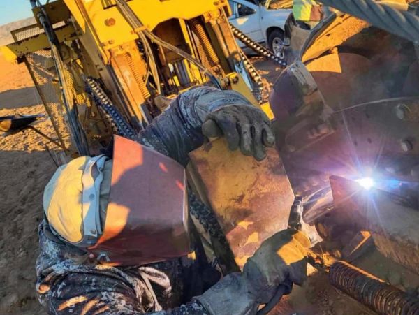 Worker welding heavy machinery with protective gear on a sunny day.