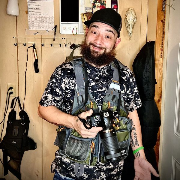 Man with a beard and cap holding a camera, dressed in camo gear.