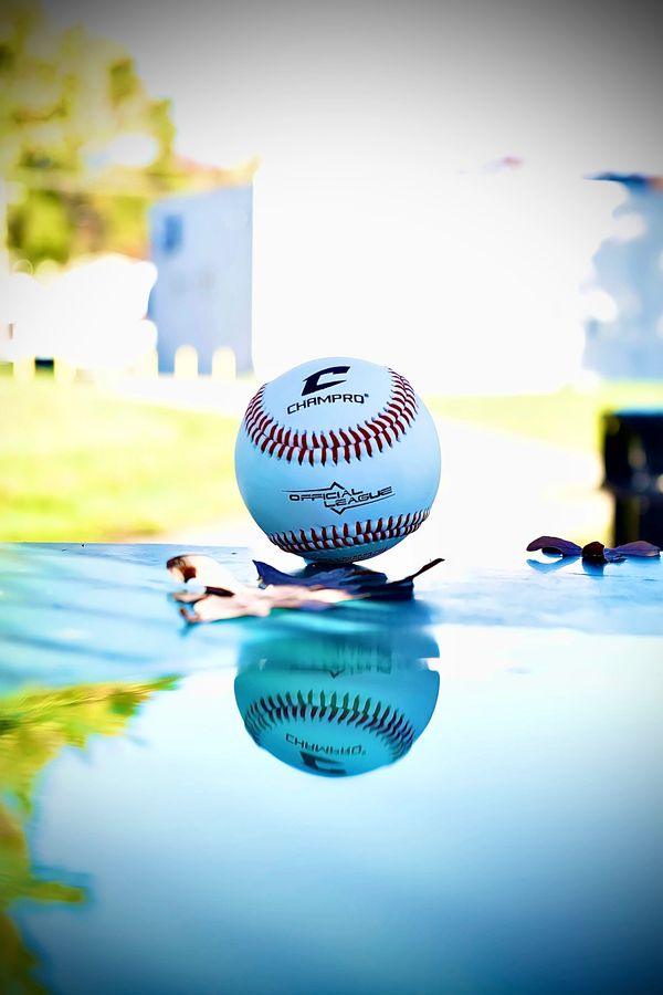 A baseball with its reflection on a shiny blue surface outdoors.