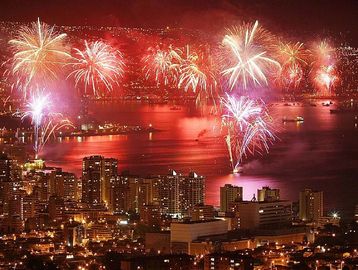 Fireworks light up the night sky over a bustling city by the water.
