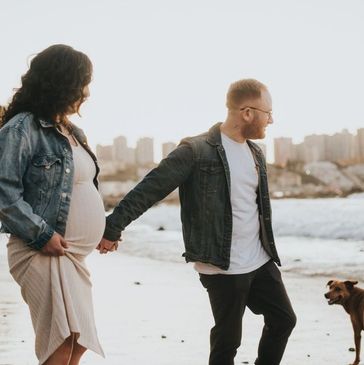 Pregnant couple holding hands on the beach with their dog at sunset.