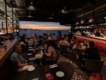 Busy restaurant with sea view during sunset and diners enjoying meals.