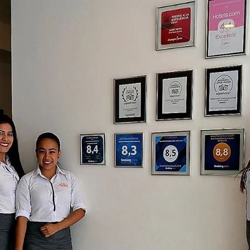 Three women in white shirts and gray skirts stand smiling in front of a wall with framed awards and ratings.
