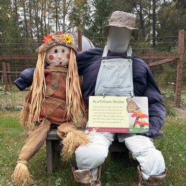 Two scarecrows sitting on a bench, one holding a pollinator sign.