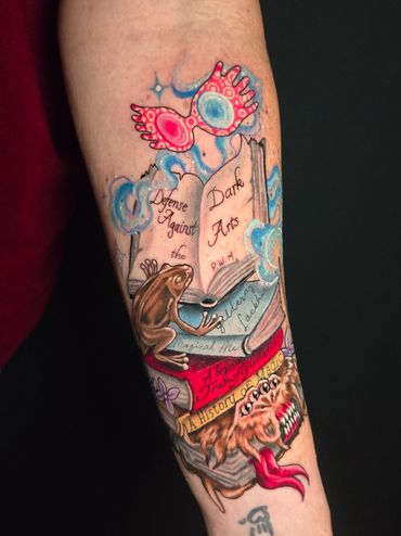 Colorful tattoo of magical books, a frog, and mystical glasses on a forearm.