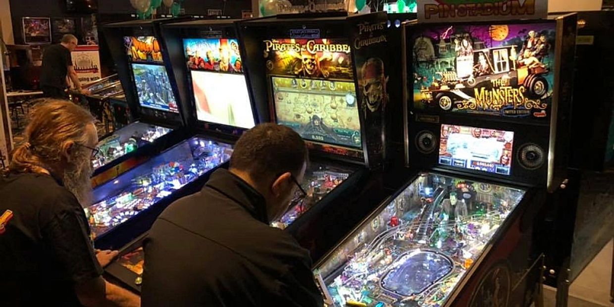 Two men playing pinball machines in a dimly lit arcade.