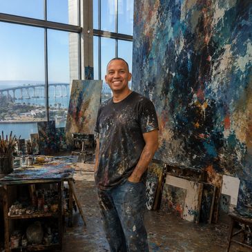 A smiling artist stands in a paint-splattered studio with large abstract paintings.
