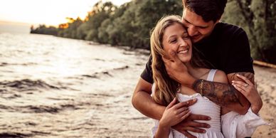 A couple embraces and smiles by the water at sunset.