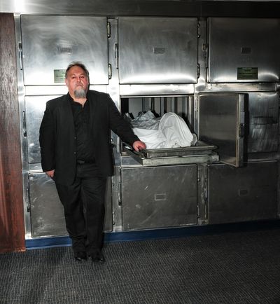 A man in black stands next to a morgue drawer with a covered body.