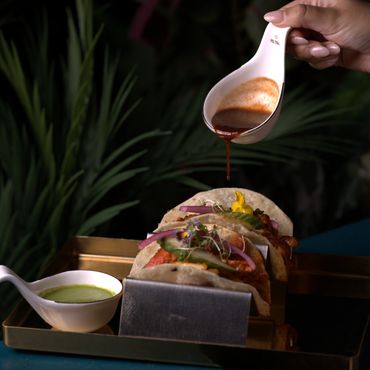 Hand pouring sauce over two tacos with dipping sauce on a tray.