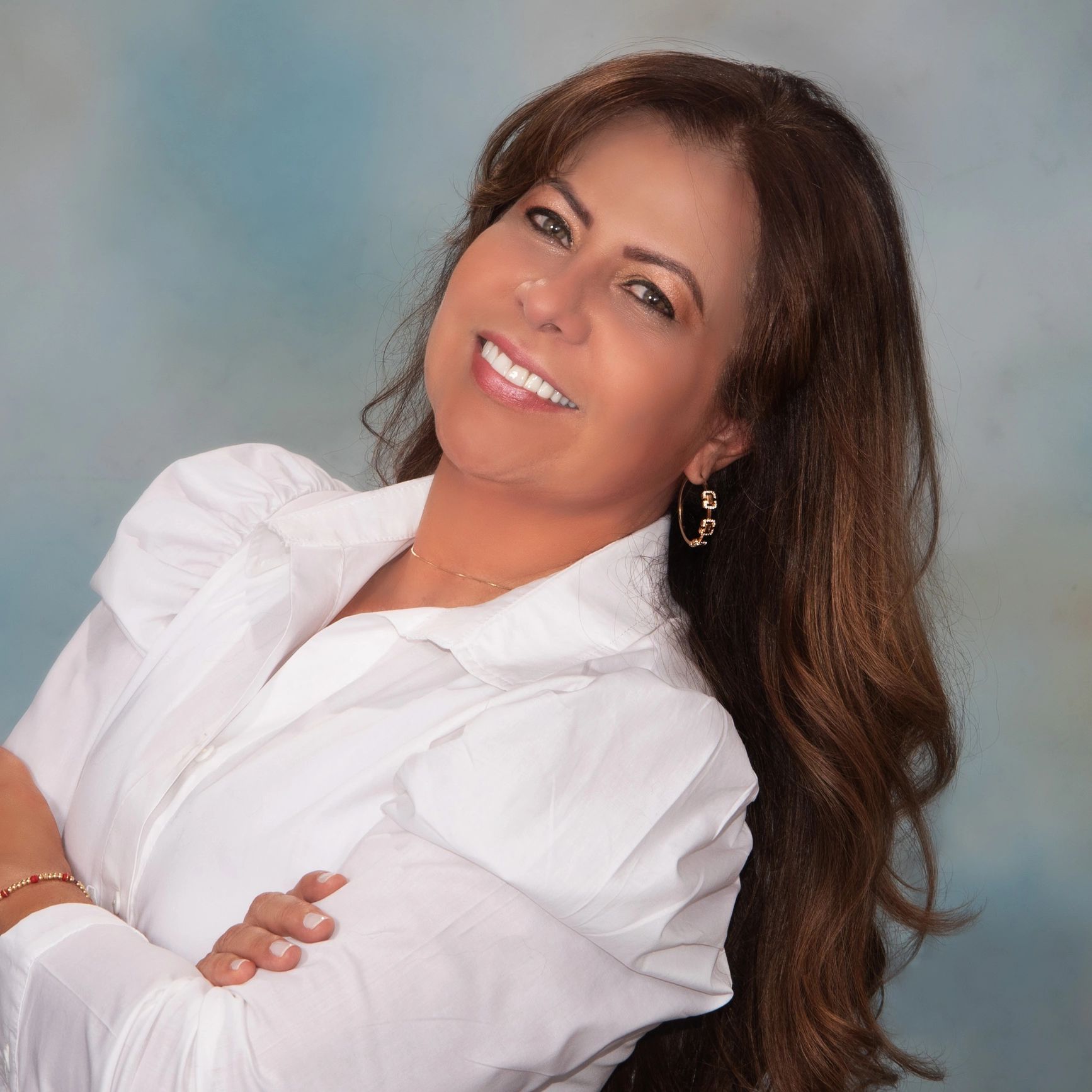 Smiling woman with long hair wearing a white blouse against a soft blue background.