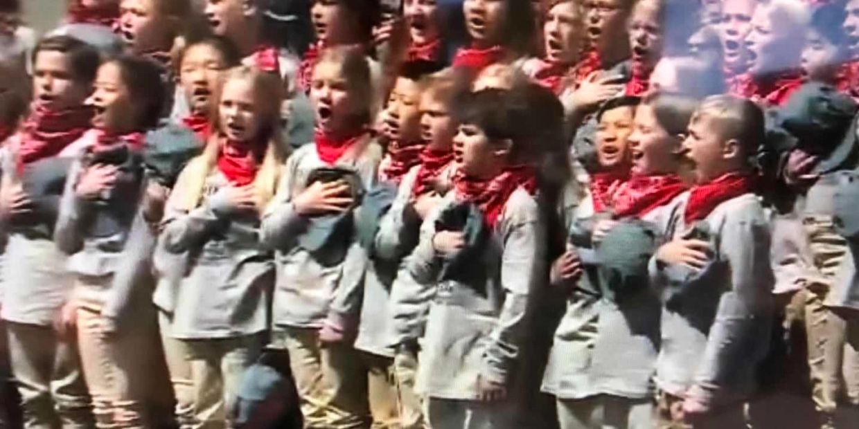 Children singing with hands on their chests wearing grey tops and red scarves.