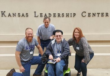 1 man in wheelchair, 2 men and 1 women standing in front of Kansas Leadership Center.