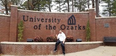 Young man sitting in front of a brick wall that says University of Ozarks