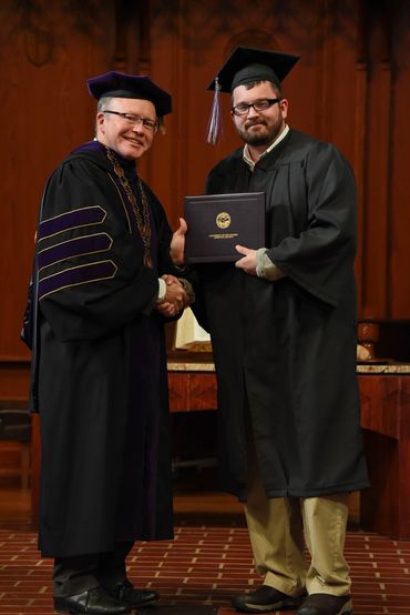 2 men standing in black graduation robes one holding a diploma.