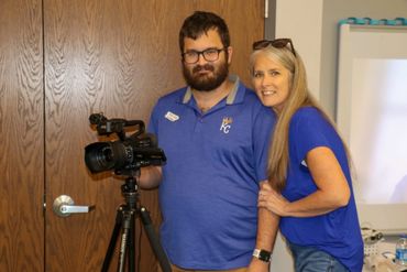 Mother and son in blue shirts standing behind a video camera.