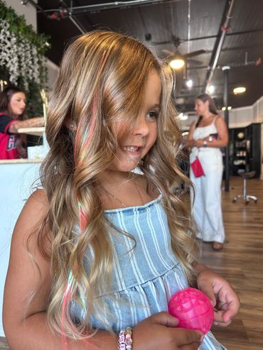 Little girl with curled hair and colorful strands holding a pink toy.