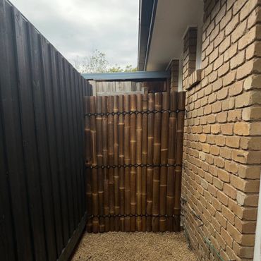 A small outdoor area with wooden decking, gravel, and a bamboo gate between brick and wooden fences.