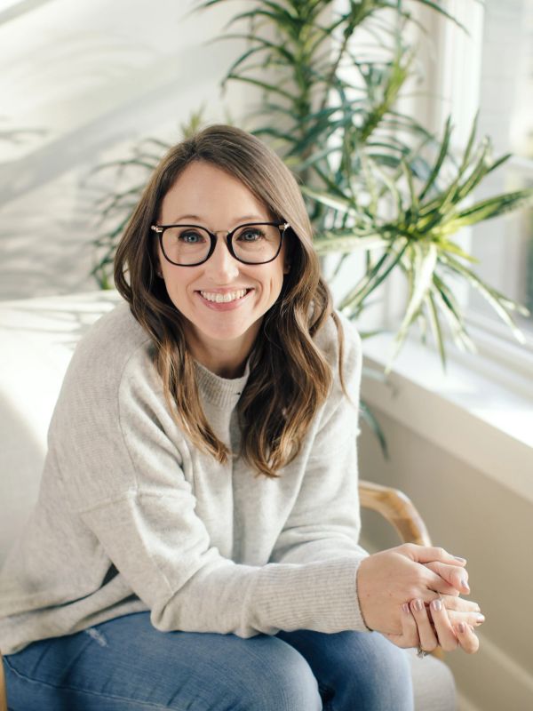 Smiling woman with glasses sitting on a chair by a sunlit window.
