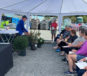 A man presents plants to a seated group under a canopy at a garden center.