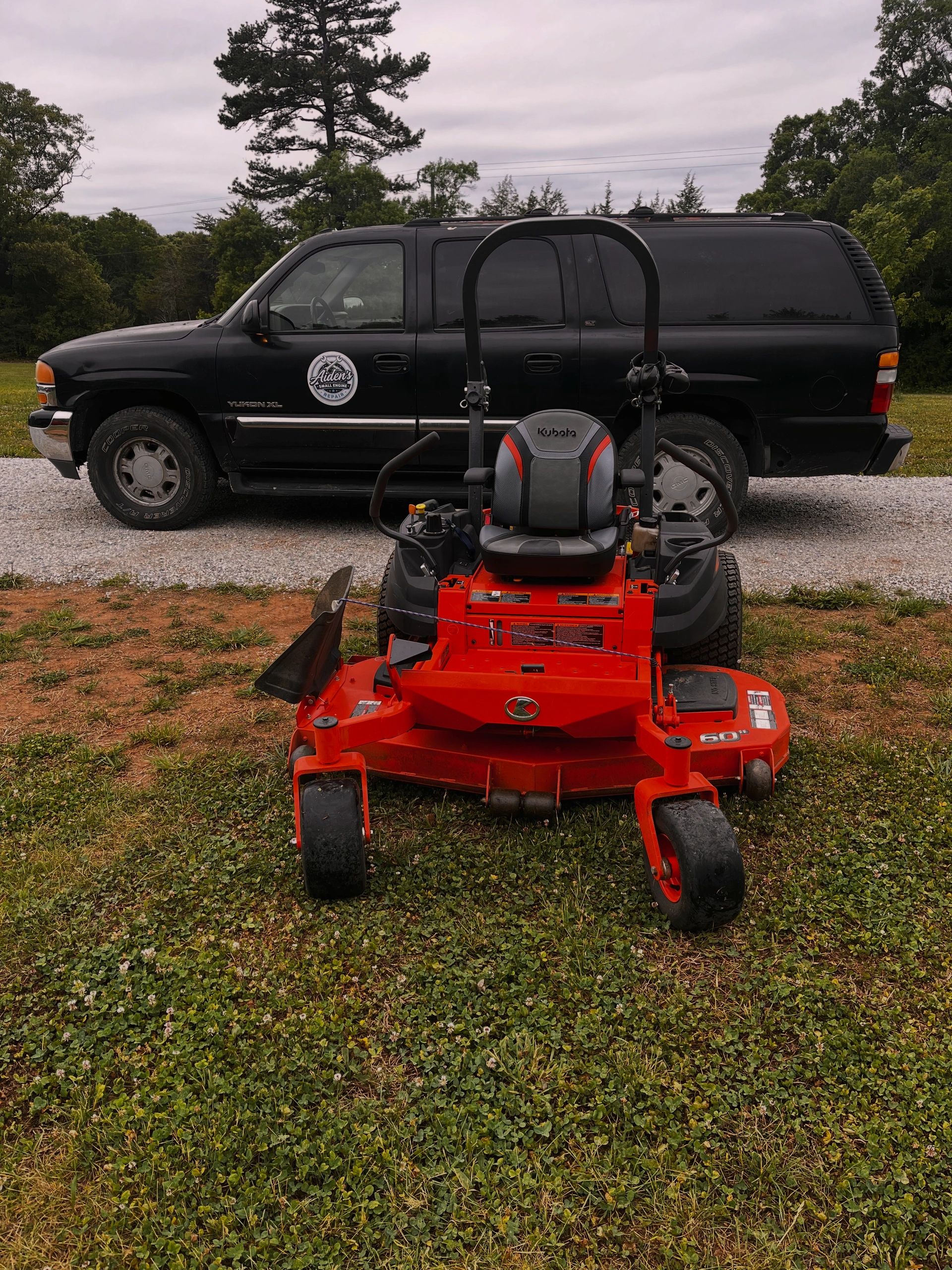 A black Yukon XL SUV and a Kubota red lawn mower on grass and gravel.