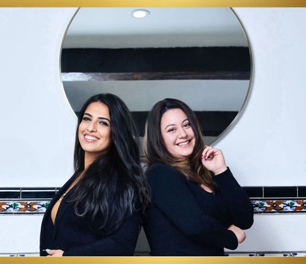 Two women in black outfits smiling confidently in front of a round mirror.