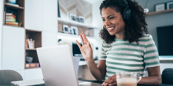 Woman with headphones video chatting on laptop, smiling and waving.