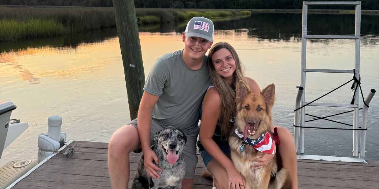 Couple with two dogs on a dock at sunset by the water.