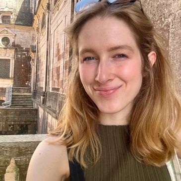 Picture of a young woman smiling softly. She has light brown hair and sunglasses on her head