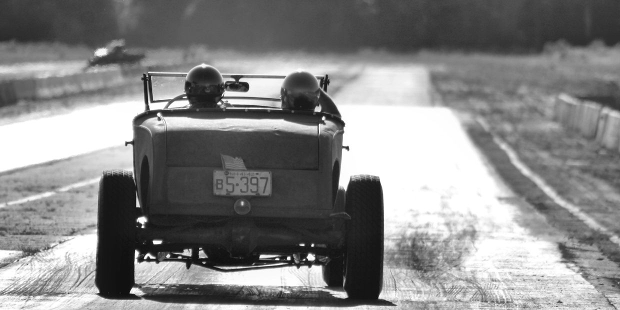 Vintage car with two passengers driving on a bright, empty road.