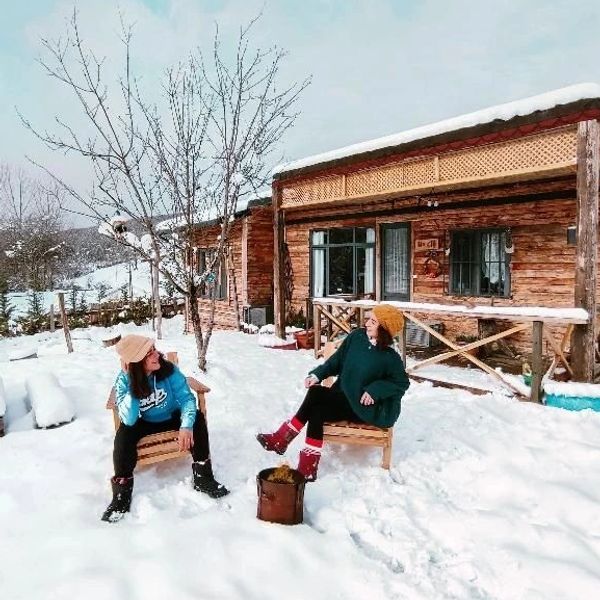 Two women sitting outside a wooden cabin in the snow, dressed warmly.