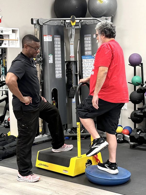 Man exercises on balance equipment with trainer's guidance.