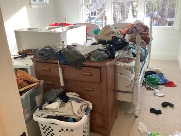Cluttered space with a bed, dresser, storage containers, and scattered clothing.