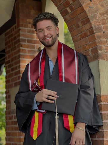 Young man in graduation gown holding cap and smiling outdoors.