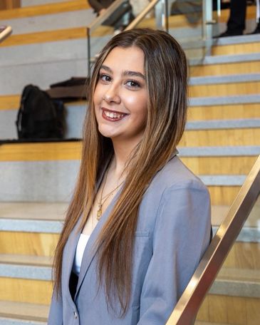 Young woman with long hair smiling, wearing a grey blazer on stairs.