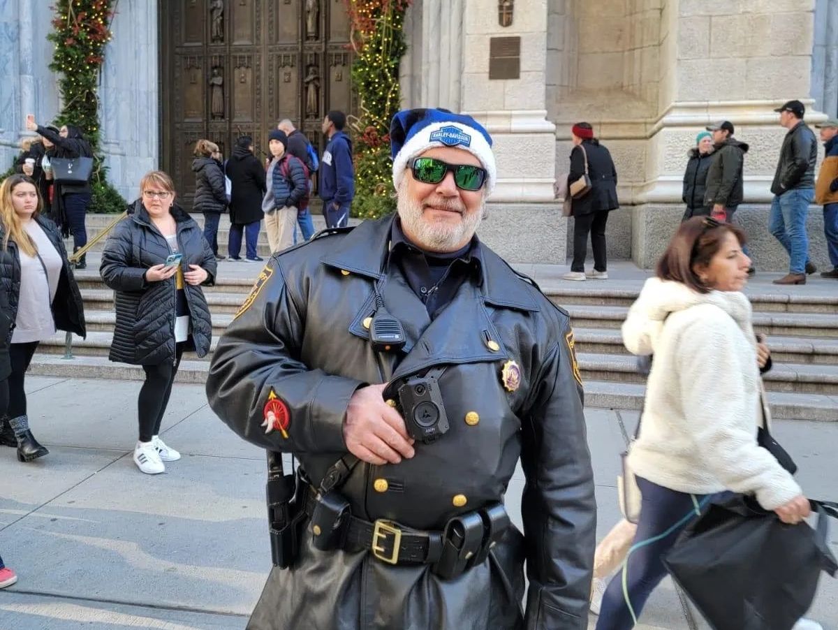 A police officer in a leather uniform and festive hat poses outdoors in front of a decorated building entrance.