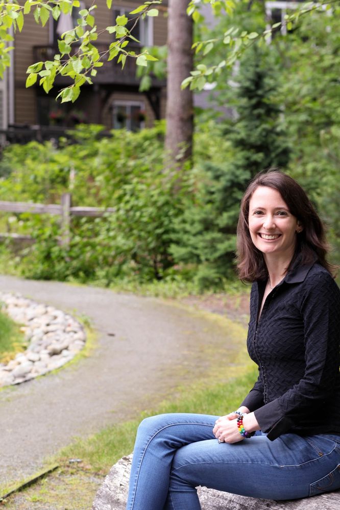 Kristiana sitting on a log smiling and facing you, along a park with a dirt path. 