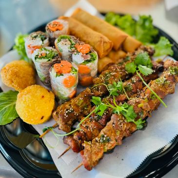 Grilled meat skewers, spring rolls, and fresh vegetable spring rolls on a black plate.