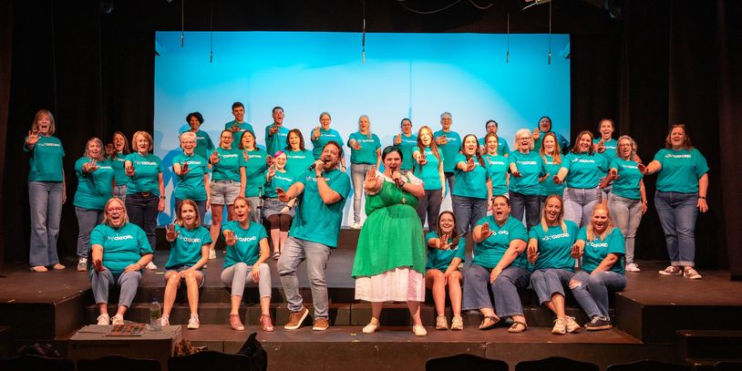 Group of people on stage pointing towards the camera, wearing teal shirts.