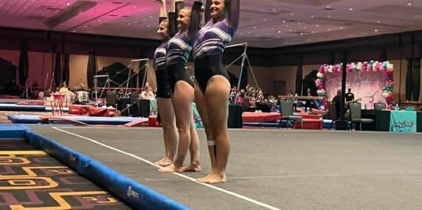 Three female gymnasts standing in a row on a gymnastics floor.