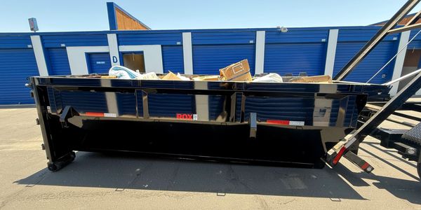 Black dumpster filled with cardboard and trash outside blue storage units.