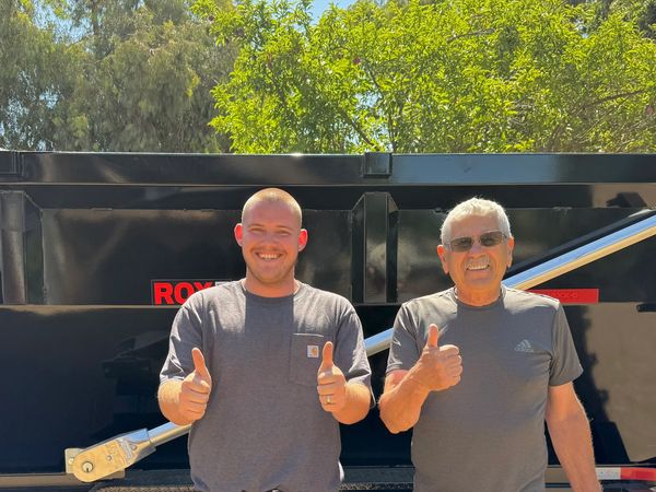 Two men standing in front of a black trailer, giving thumbs up.