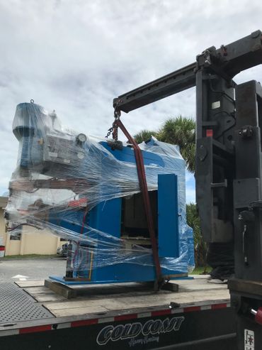 Large blue industrial machine being lifted by a crane on a flatbed truck.